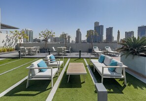 A rooftop terrace with white sofas and tables, arranged on artificial grass, overlooking a cityscape with modern buildings. - Modern Studio in Oxford Boulevard JVC (Dubai)