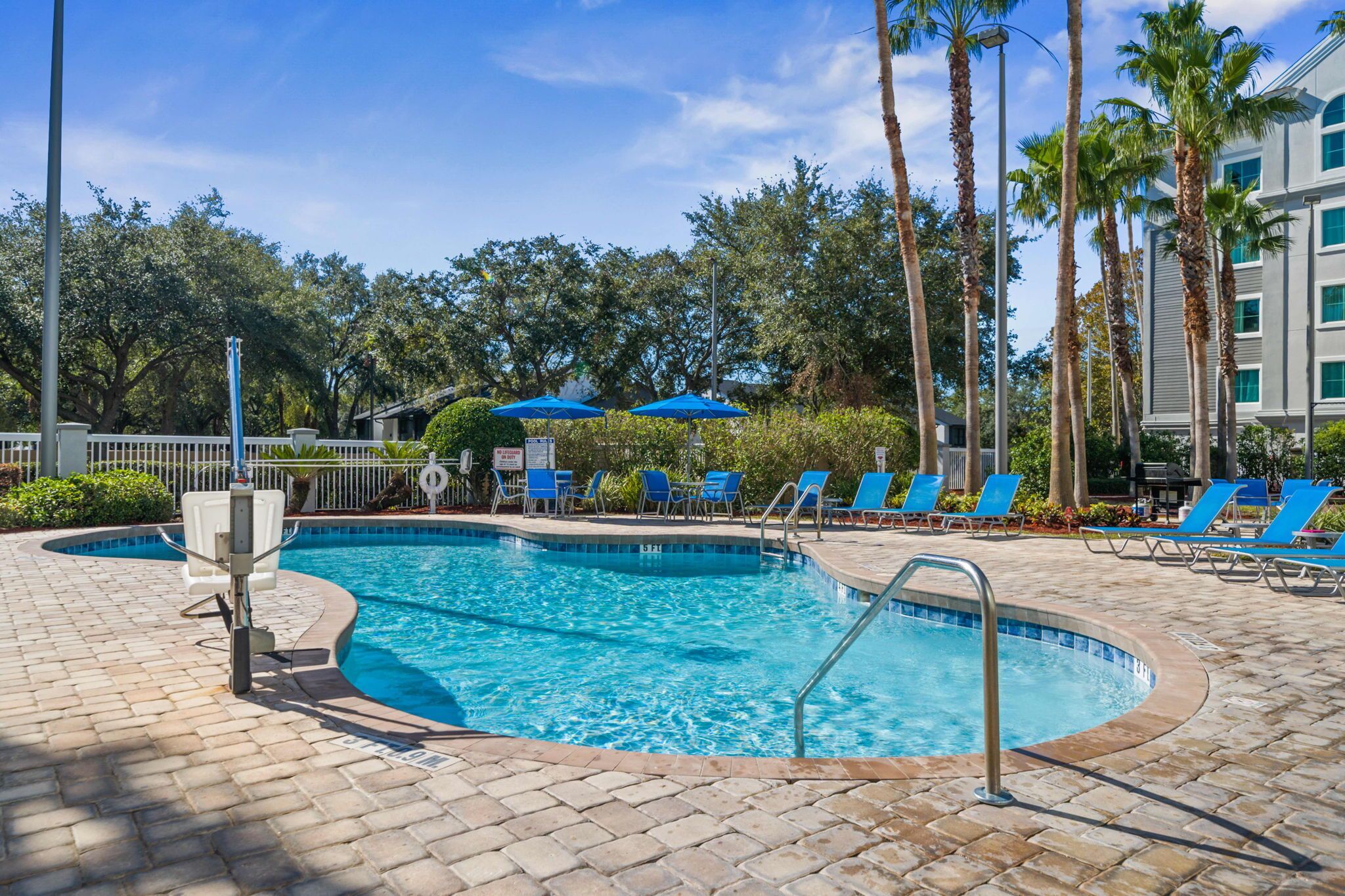 A swimming pool with blue chairs and umbrellas, surrounded by palm trees and a building in the background.