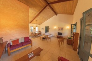 A spacious living area with a high ceiling, wooden furniture, a sofa, a dining table, and a television.