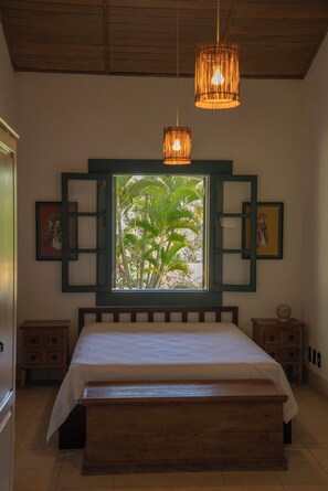 A bedroom with a bed, wooden headboard, two bedside tables, a window with a view of palm trees, and a hanging light fixture.