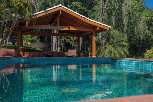 A clear swimming pool with a wooden deck and lounge chairs, surrounded by lush greenery.