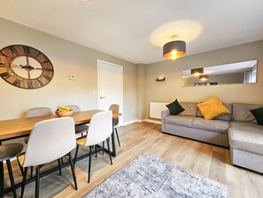 Una sala moderna con comedor, un sofá gris con cojines amarillos y verdes, una mesa de comedor de madera con sillas blancas y un reloj de pared grande.