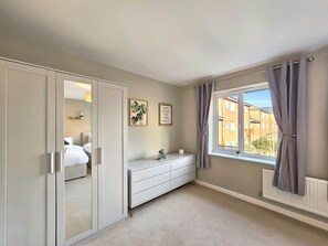 A bedroom with a large wardrobe, a bedside table, a window with curtains, and a view of buildings outside. - The Cobblestone Townhouse by Fortuna Property Portfolio (Cheltenham)