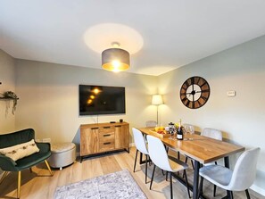 Una sala moderna con una mesa de comedor de madera, sillas blancas, un sillón verde, un televisor y un reloj en la pared.