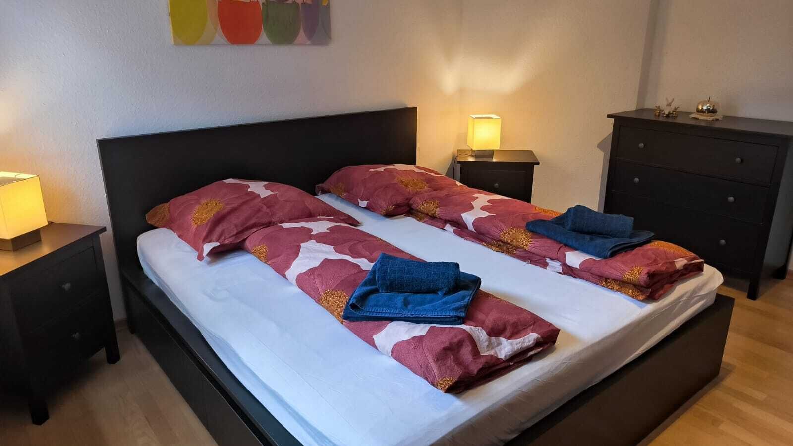 A double bed with a white and red floral bedspread, two pillows, and folded blue towels. There are two bedside tables with lamps, a dark dresser, and a wall with a colorful abstract painting.