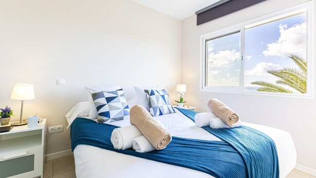 Una cama bien hecha con ropa de cama azul y blanca, dos toallas enrolladas y almohadas estampadas con diseños geométricos.