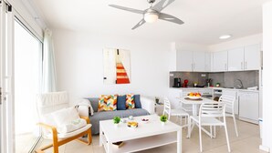 Una sala de estar moderna con sofá, mesa de centro y sillas, un comedor con mesa y sillas, y una cocina con gabinetes y electrodomésticos blancos.