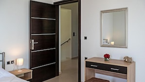 Habitación de hotel con cama, un escritorio de madera con cajones, un espejo, una mesita de noche con lámpara y una puerta que da a un pasillo.