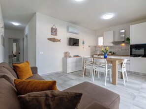 Ein modernes Wohnzimmer mit einem braunen Sofa, einem Esstisch mit weißen Stühlen und einer Küchenzeile mit weißen Schränken und einem schwarzen Backofen.
