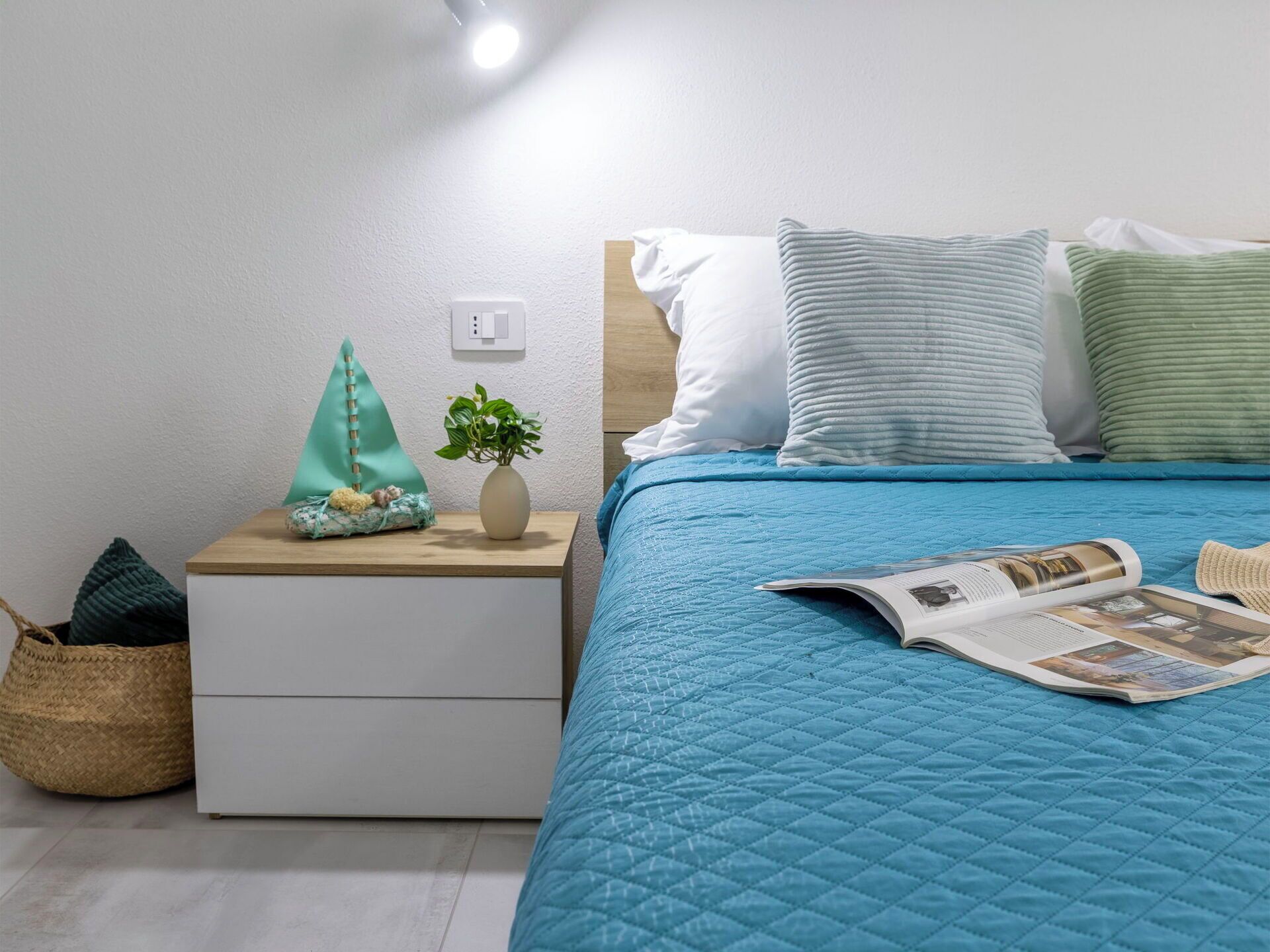 Ein ordentlich bezogenes Bett mit einer blauen Stegdecke, ein Nachttisch mit einer Pflanze und eine Zeitschrift auf dem Bett.