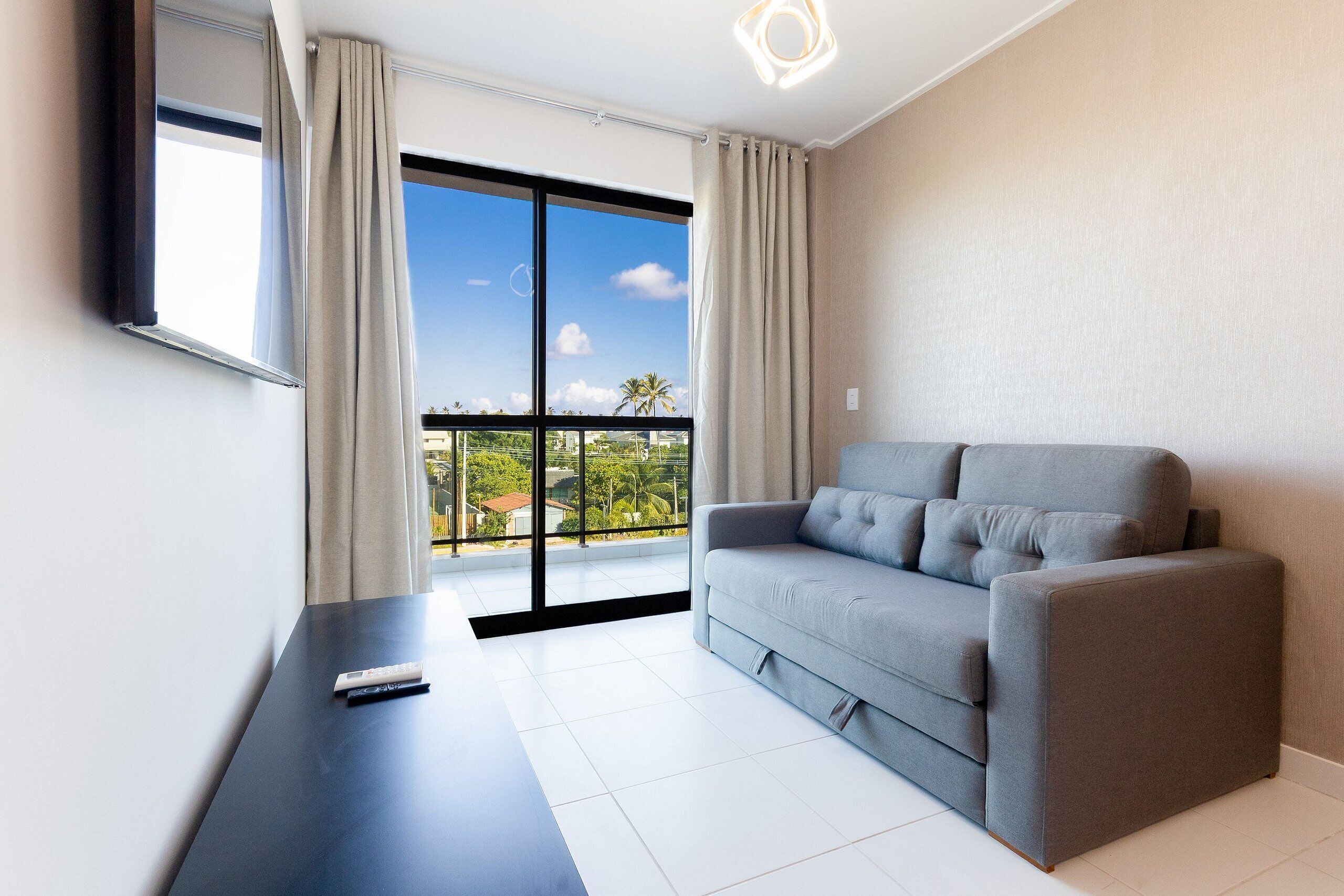 Una sala de estar moderna con un sofá gris, un televisor de pantalla plana montado en la pared, un ventanal con cortinas y un balcón con vistas a palmeras.