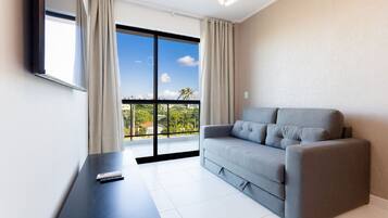 Una sala de estar moderna con un sofá gris, un televisor de pantalla plana montado en la pared, un ventanal con cortinas y un balcón con vistas a palmeras.