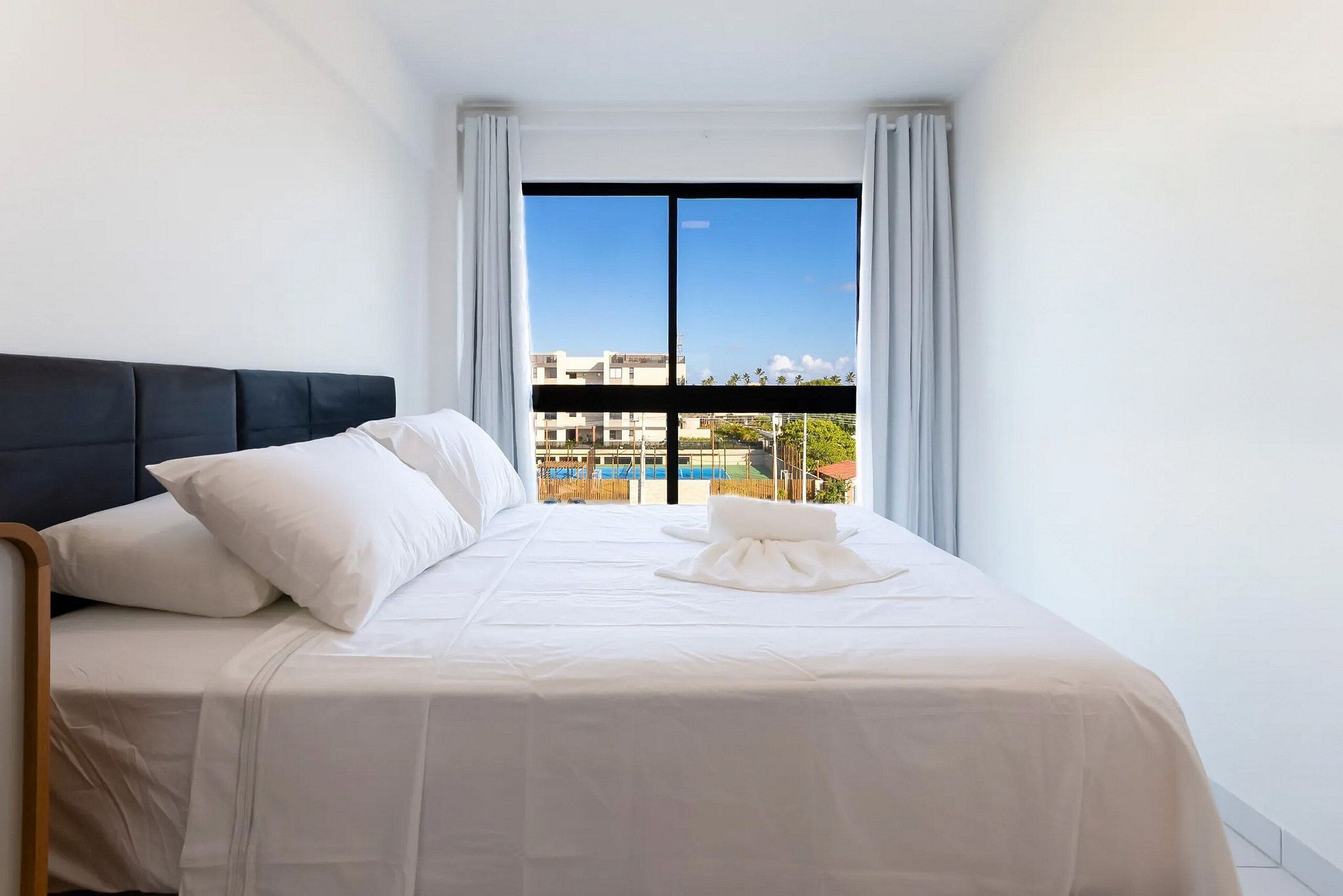Quarto de hotel com cama, roupa de cama branca, cabeceira preta e vista para edifícios e piscina através de uma grande janela.