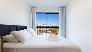 A hotel room with a bed, white bedding, a black headboard, and a view of buildings and a pool through a large window. - Malia Beach #a301 - Resort por Carpediem (Ipojuca)
