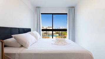 Habitación de hotel con cama, ropa de cama blanca, cabecera negra y vistas a edificios y una piscina a través de un ventanal grande.