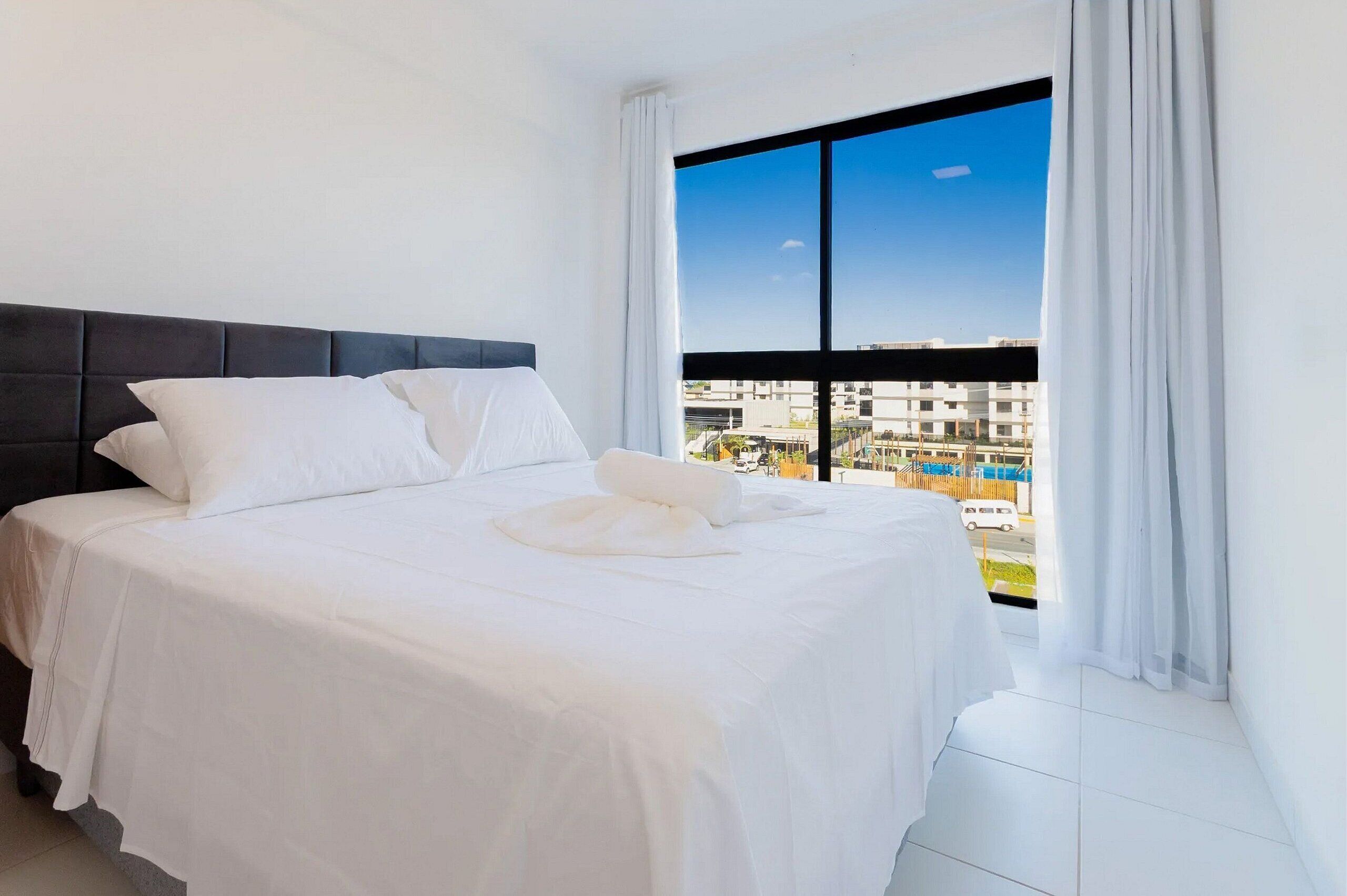 A hotel room with a large bed, a dark headboard, white bedding, and a view of buildings and a pool through a large window.