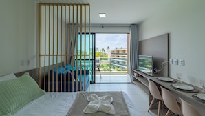 Habitación de hotel con cama, escritorio con televisor y balcón con vistas a edificios y vegetación.