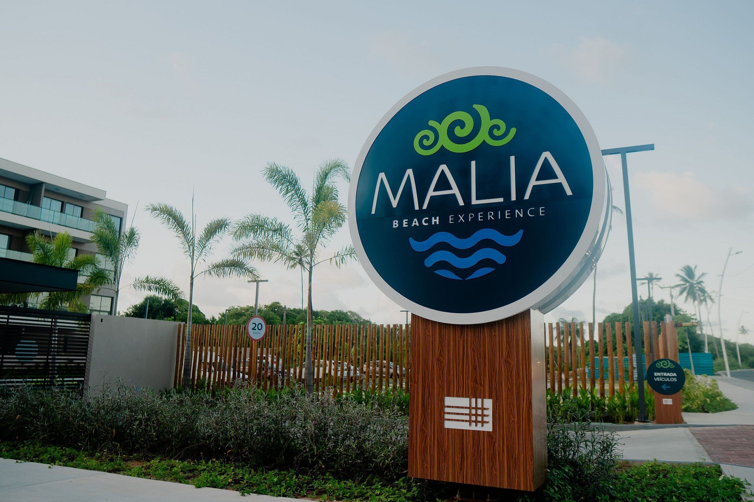 Placa para 'MALIA Beach Experience' com um logotipo, rodeada por vegetação e um cercado.