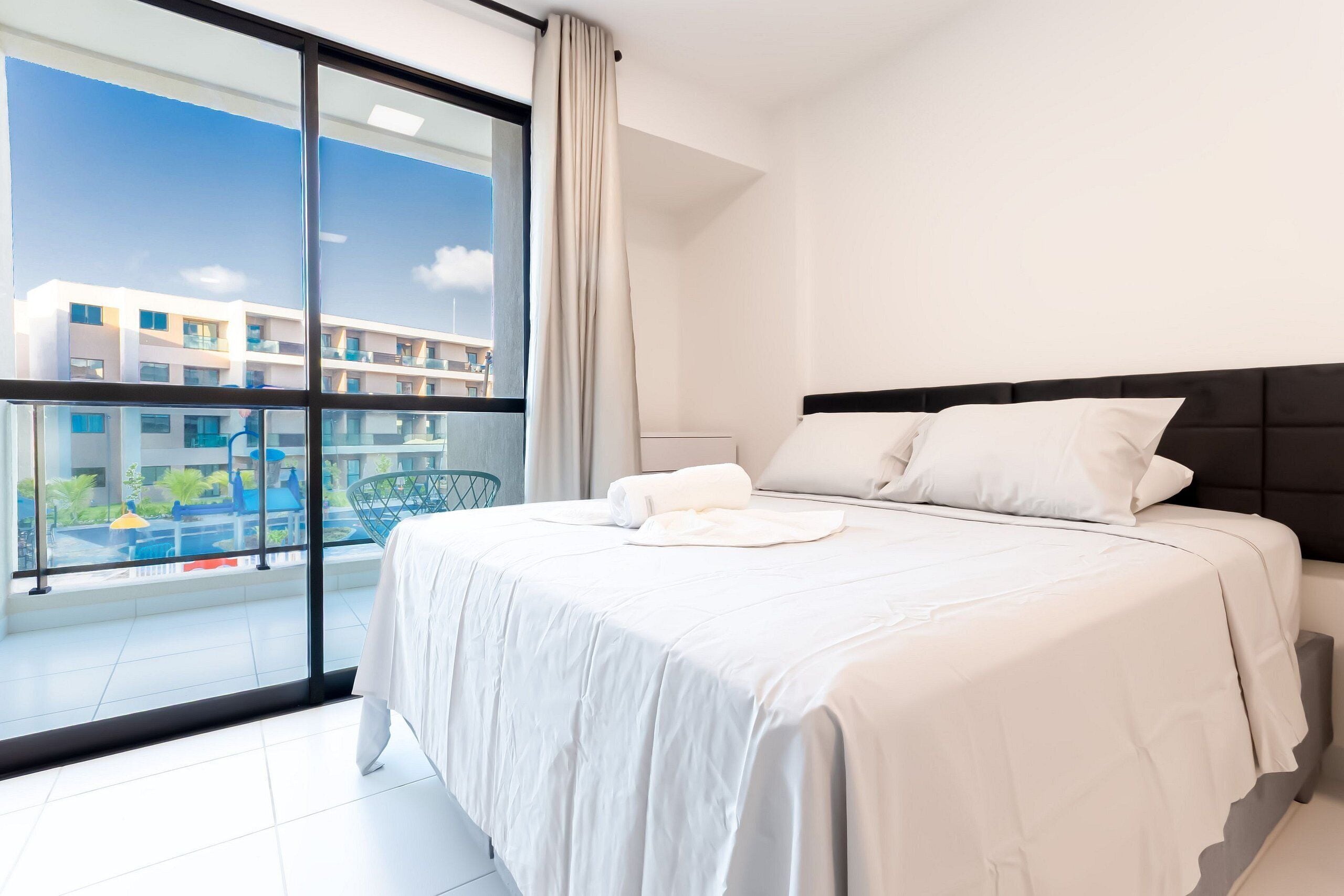 Quarto de hotel com uma cama grande, vista para uma piscina e edifícios, e varanda com porta de vidro.