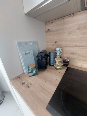 Eine Küchenarbeitsplatte mit einer Kaffeemaschine, einem Glas mit Korkverschluss, einem schwarzen Behälter und einem Metallregal mit Tassen.