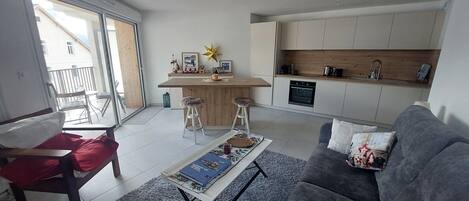 A modern living room with a grey sofa, a coffee table with books, and a kitchen area with wooden cabinets and a built-in oven.