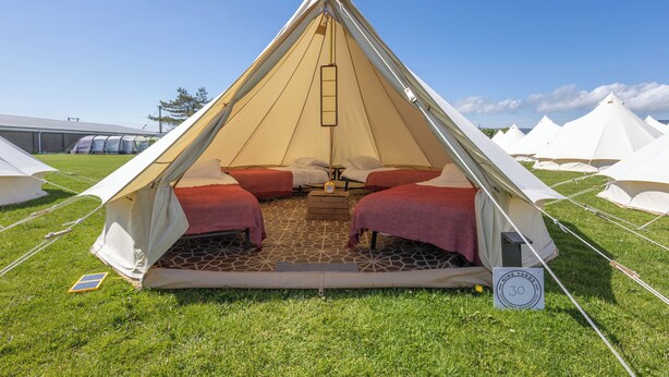 Une grande tente en forme de pyramide, dotée de deux lits et d’une table centrale, installée sur un champ herbeux, avec d’autres tentes en arrière-plan.