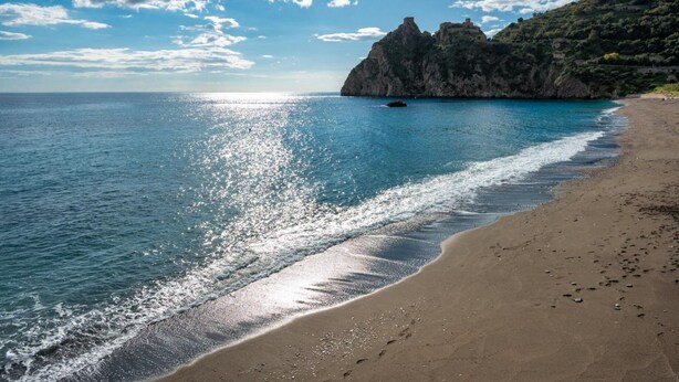 Ein Strand mit klarem, blauem Wasser, einem sandigen Ufer und felsigen Klippen im Hintergrund.