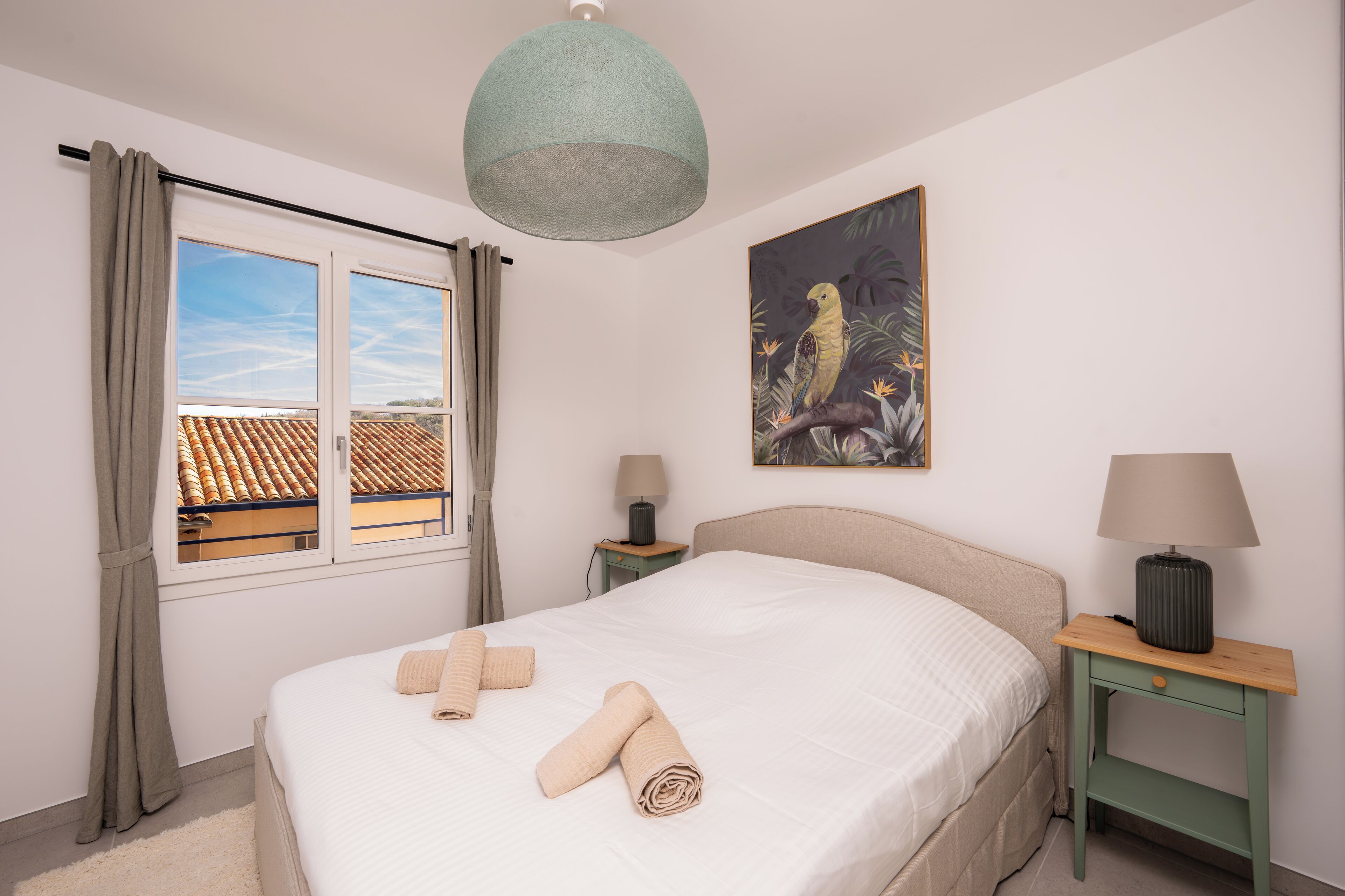 A neatly made bed with rolled towels, a painting of a bird, a lamp, and a view of a building through a window with curtains.