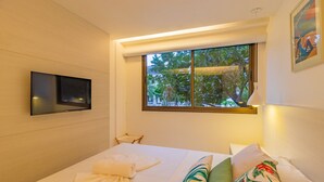 A hotel room with a bed, a TV mounted on the wall, a window with a view of greenery, and a painting on the wall. - Malawi #104a - Seaside Apartment by Carpediem (Ipojuca)