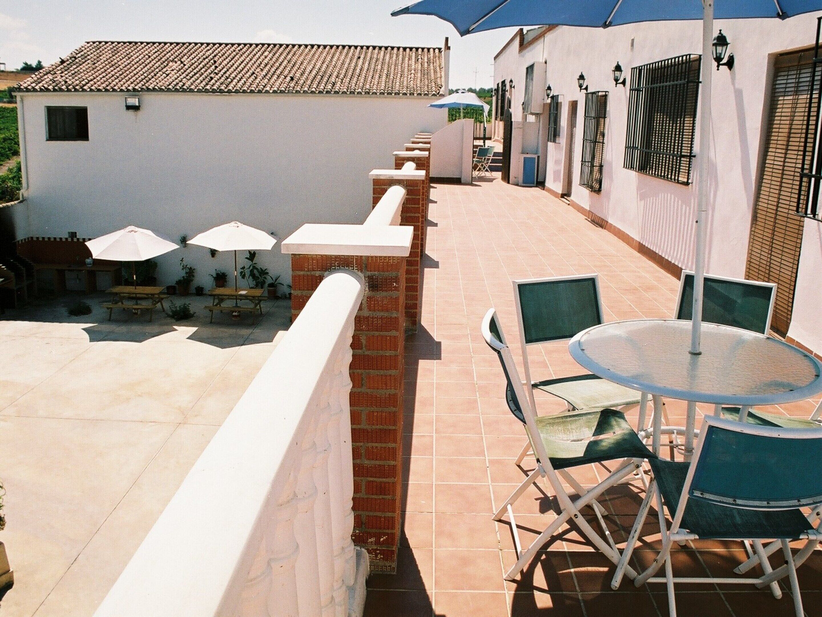 A patio area with a round table and chairs, a white wall with a window, and a tiled roof.