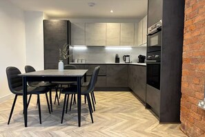 A modern kitchen with dark cabinetry, a dining table with chairs, and a brick accent wall. - Manchester Apt, Free Parking, Couples & Families near Etihad & Co-Op Live (Manchester)