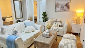 A living room with a white sofa, a wicker coffee table, and a matching ottoman. There is a mirror on the wall, a plant, and a lamp on a side table.