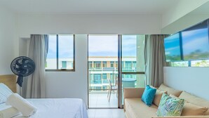Et hotelværelse med seng, sofa, fjernsyn, ventilator og balkon med udsigt til bygninger og en palme.