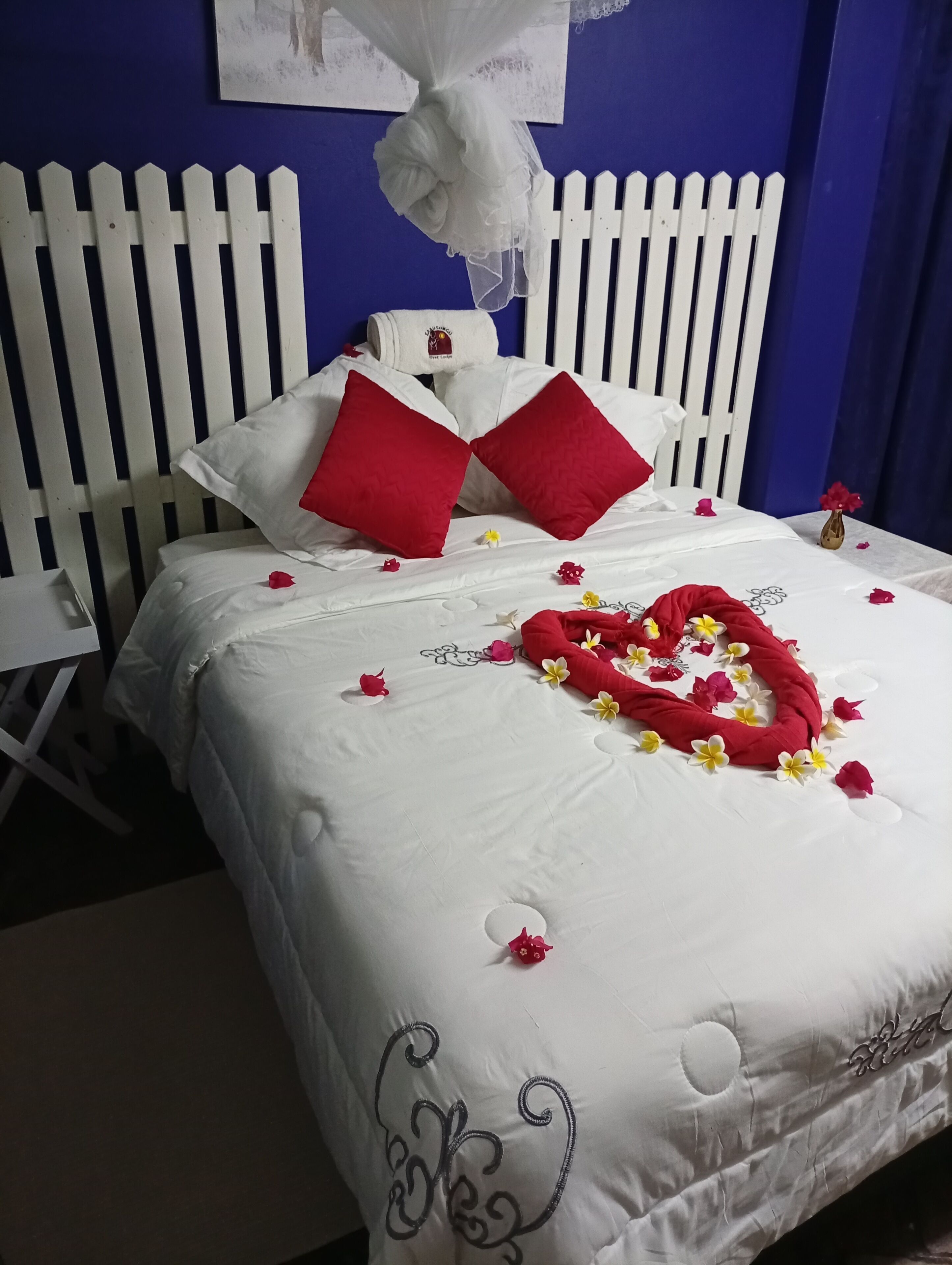 A neatly made bed with white and red bedding, a white headboard, and a decorative heart made of flowers on the bed.