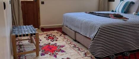 Un dormitorio con un ventanal grande, una cama con ropa de cama a rayas, una silla de madera y una alfombra estampada.