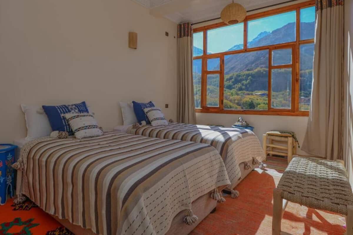 Un dormitorio con dos camas, una ventana con vista a la montaña, un taburete de madera pequeño y una alfombra roja.