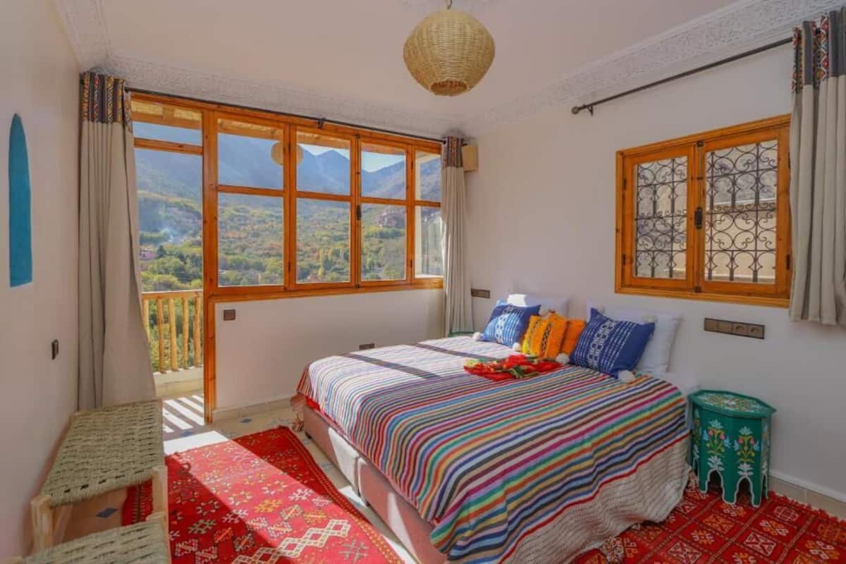 Un dormitorio con un ventanal que ofrece vistas a la montaña, una cama con ropa de cama de colores y un taburete pequeño verde.
