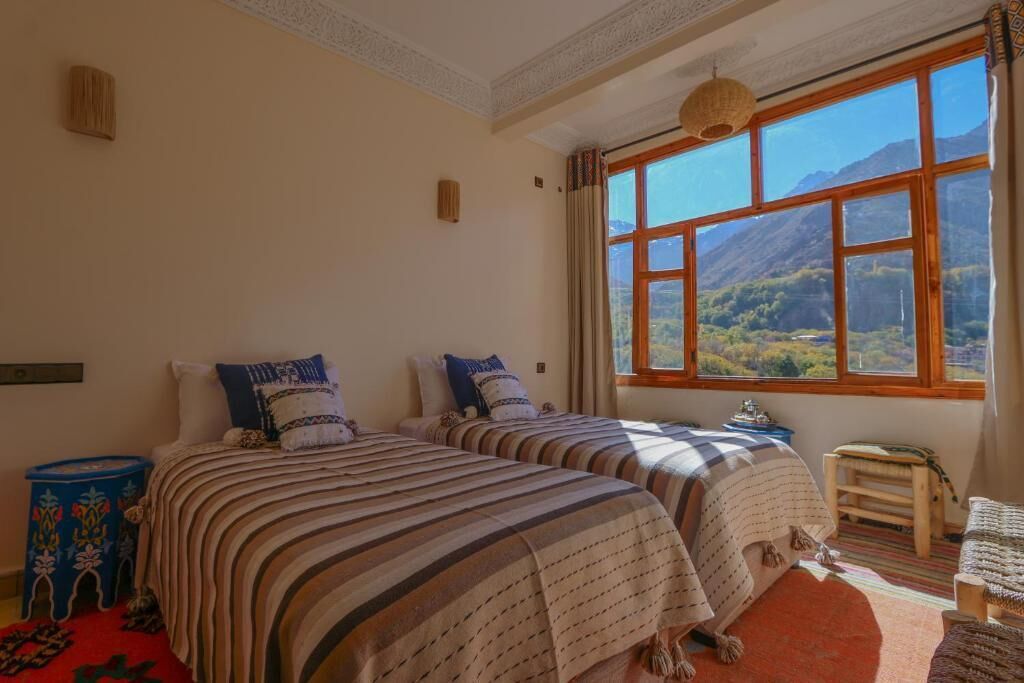 Une chambre avec deux lits, un petit tabouret et une vue sur les montagnes, visible à travers de grandes fenêtres.