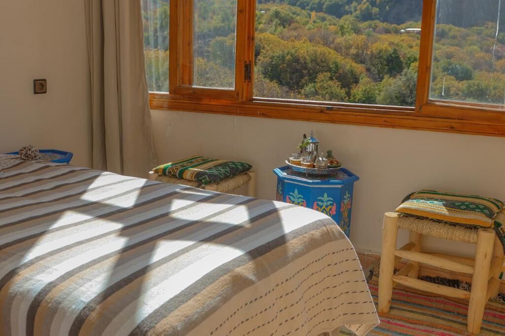 Un dormitorio con una cama, un taburete pequeño, un armario azul y una ventana con vistas a un bosque.