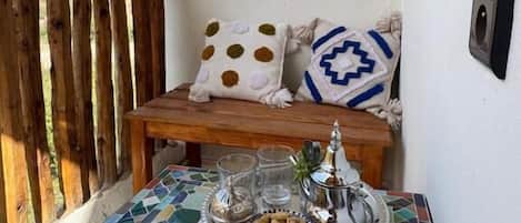 A balcony with a wooden bench, a tiled table with a tray of nuts and a silver container, and two pillows with distinct patterns.