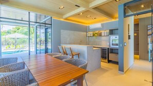 A modern kitchen with a central island, a dining area with wicker chairs, and large windows offering a view of a pool and greenery. - Cais Eco #310 - Ape com Piscina por Carpediem (Ipojuca)