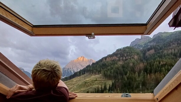 Una persona apoyada en el marco de madera de una ventana, con vistas a un paisaje montañoso.
