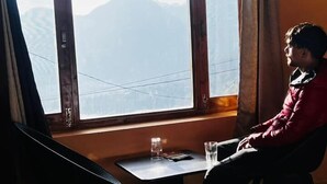 Ein Zimmer mit großem Fenster und Blick auf die Berge, ein kleiner Tisch mit einem Glas und einem Buch sowie ein Bett mit weißer Bettdecke.