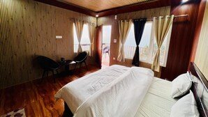 Ein Schlafzimmer mit einem großen Bett, einem Schreibtisch und einem Stuhl. Vor den Fenstern hängen Vorhänge, und durch die Tür ist ein Balkon zu sehen.