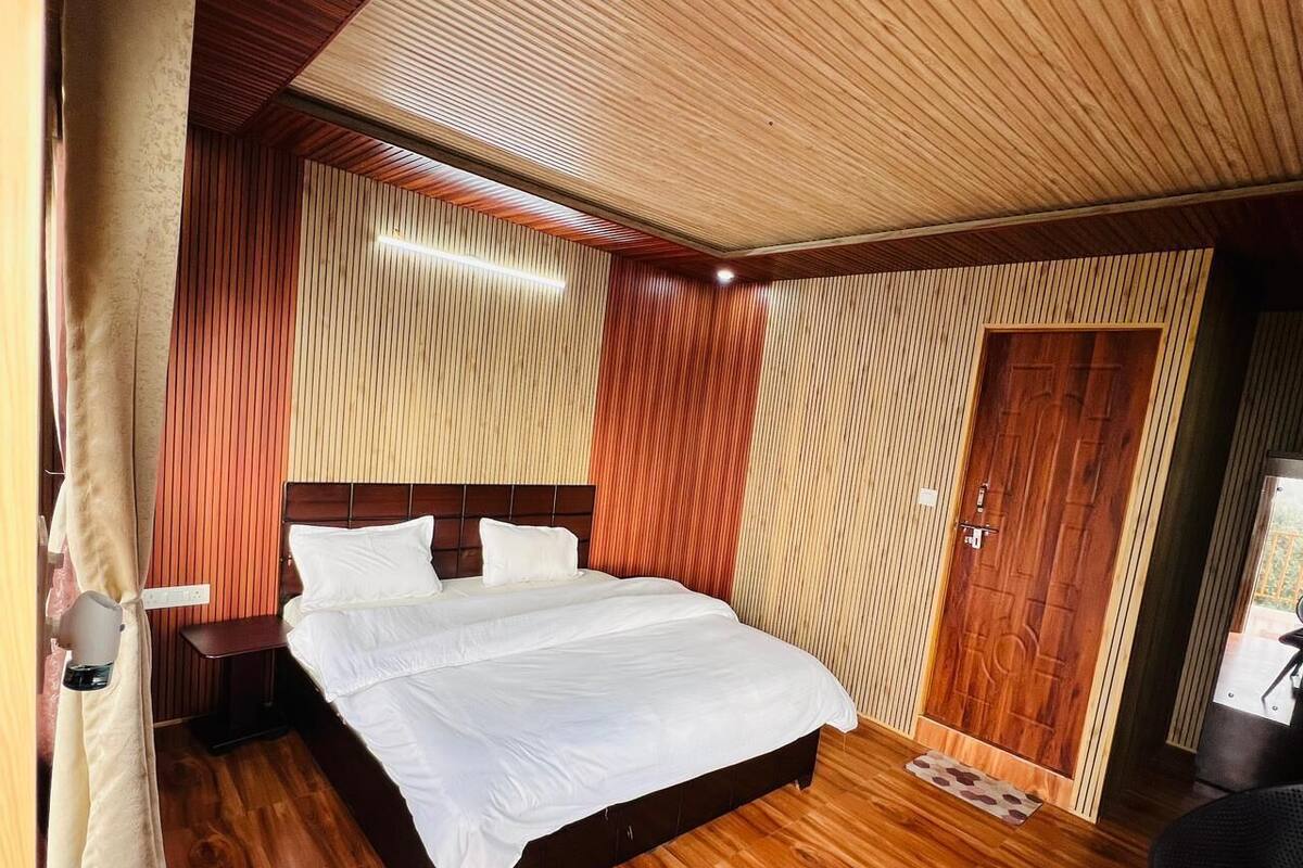 A bedroom with a wooden floor, a bed with white bedding, a wooden door, and a nightstand with a lamp.