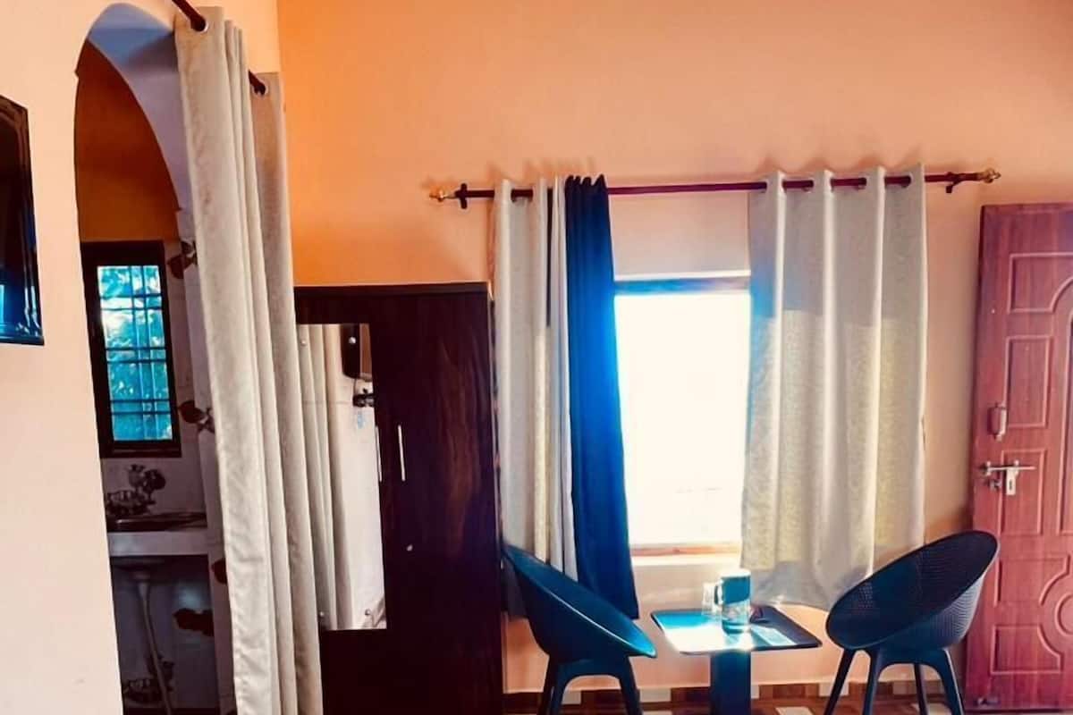 A hotel room with a wooden parquet floor, two chairs, a bed, a desk with a chair, a window with curtains, and a door.