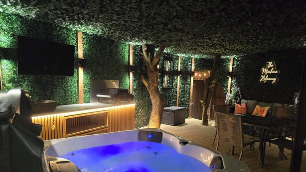A hot tub with a digital control panel, a wooden deck, a bar area with a TV, and a seating area with a neon sign.