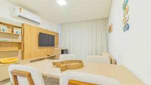 A modern room with a flat-screen TV mounted on a wooden panel, a dining table with chairs, and a large window with curtains. - Cais Eco #212 - Apartment on the Sand by Carpediem (Ipojuca)