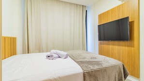A hotel room with a bed, a television mounted on the wall, and a window with curtains. - Cais Eco #212 - Apartment on the Sand by Carpediem (Ipojuca)