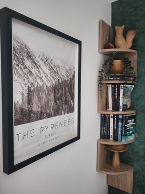 Un tableau encadré représentant « les Pyrénées » est accroché au mur, ainsi qu’une bibliothèque en bois remplie de livres et une petite plante, et un mur vert à texture.